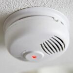 Round First Alert smoke alarm on a clean white ceiling with a small red LED indicator illuminated