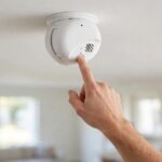 A person's hand pressing the Test/Silence button on a First Alert smoke alarm mounted on a white ceiling